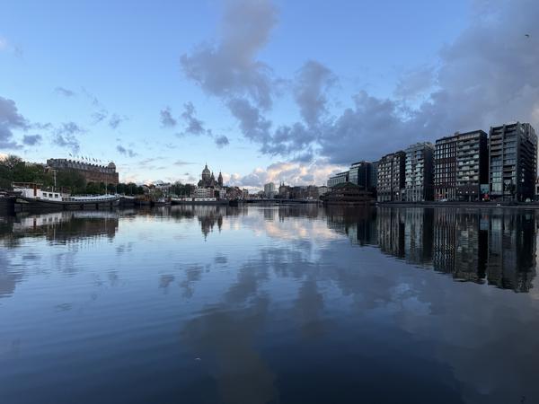 This tranquil evening scene captures the Prins Hendrikkade in Centrum, Amsterdam, showcasing a wide expanse of calm water reflecting the dramatic sky and city architecture